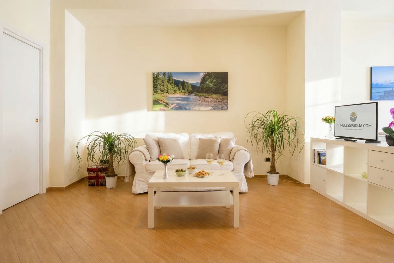 Ein Wohnzimmer mit einem weißen Sofa, einem Couchtisch, zwei Blumentöpfen, einem Gemälde eines Flusses und einem Fernseher auf einer weißen Kommode.