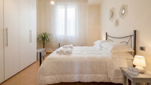 A hotel room with a bed, a bedside table with a lamp, a potted plant, and a window with sheer curtains.