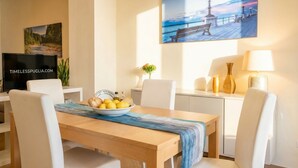 A dining area with a wooden table, white chairs, a bowl of fruit, a television, and a painting of a waterfront scene.