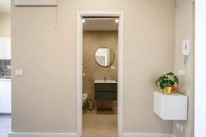 A narrow hallway leading to a bathroom with a sink, mirror, and toilet. A small plant is on a shelf to the right.