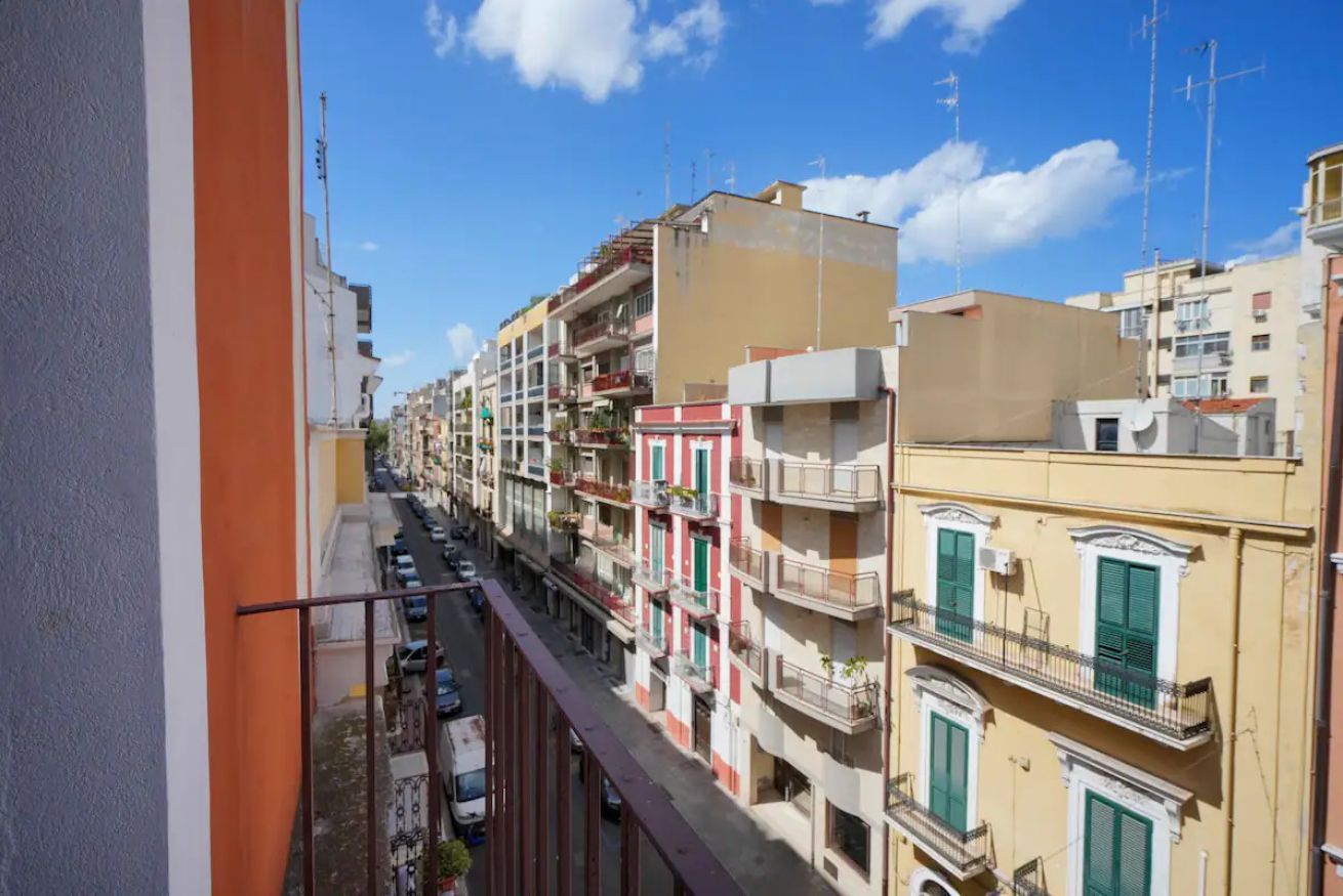 A row of multi-story buildings with balconies, a clear sky, and a street with parked cars.