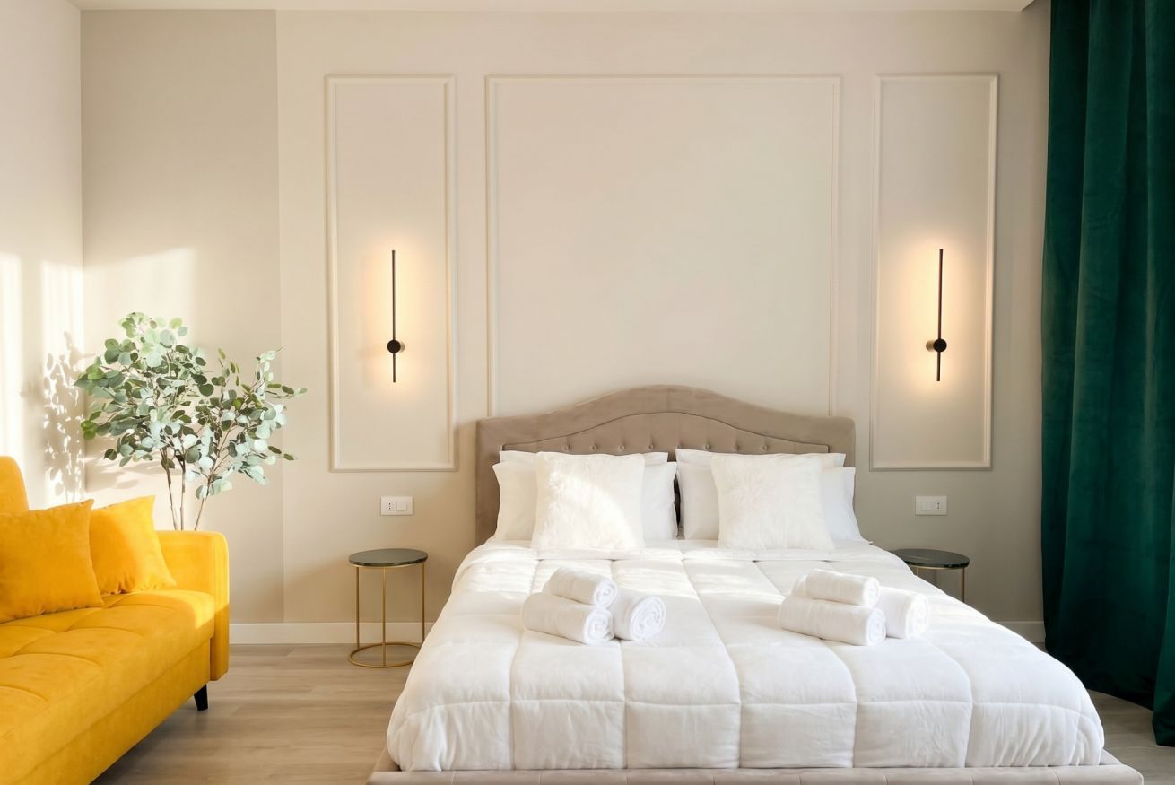 Un dormitorio ordenado con una cama, dos mesitas de noche, un florero y un sofá amarillo.