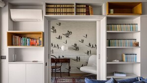 Een moderne woonkamer met een witte bank, een houten salontafel en een kamerkleed met een patroon. Er staan boekenkasten vol boeken en een muur met een muurschildering.