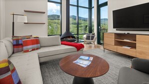A modern living room with a sofa, a coffee table, a fireplace, a TV, and large windows offering a view of a forested landscape.