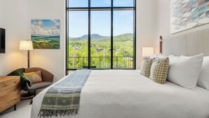 A bedroom with a large window, a bed, a chair, a nightstand, and a painting.