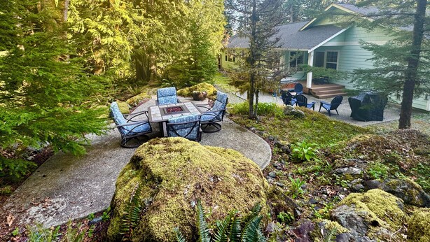 A cozy outdoor seating area with a table and chairs, surrounded by lush greenery and a house in the background.