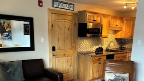 Una sala de estar acogedora con un sofá de cuero marrón, un tapete de cuadros y un sillón de cuero marrón. Una puerta de madera da a una cocina con armarios de madera y un salpicadero de azulejos.