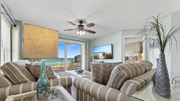 A living room with striped sofas, a glass coffee table, a ceiling fan, and a view of the beach through large windows.