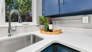 Une cuisine avec un évier en acier inoxydable, une fenêtre donnant sur des arbres, un meuble bleu et un plateau avec une bouteille et des verres.