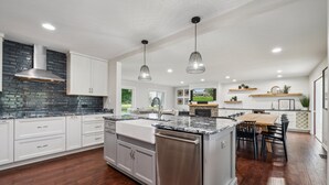 A modern kitchen with white cabinets, a stainless steel dishwasher, and a large island with a sink. There are pendant lights hanging above the island and a large window providing natural light.