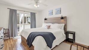 A bedroom with a bed, a ceiling fan, a nightstand, a chair, and a view of a balcony.