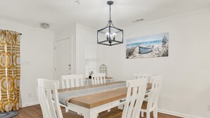 A dining area with a wooden table, white chairs, a patterned curtain, and a painting of a beach scene.