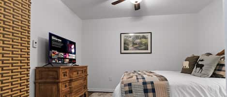 Un dormitorio con una cama, un aparador de madera, un ventilador de techo, un cuadro enmarcado en la pared y un televisor que muestra un cartel de película.
