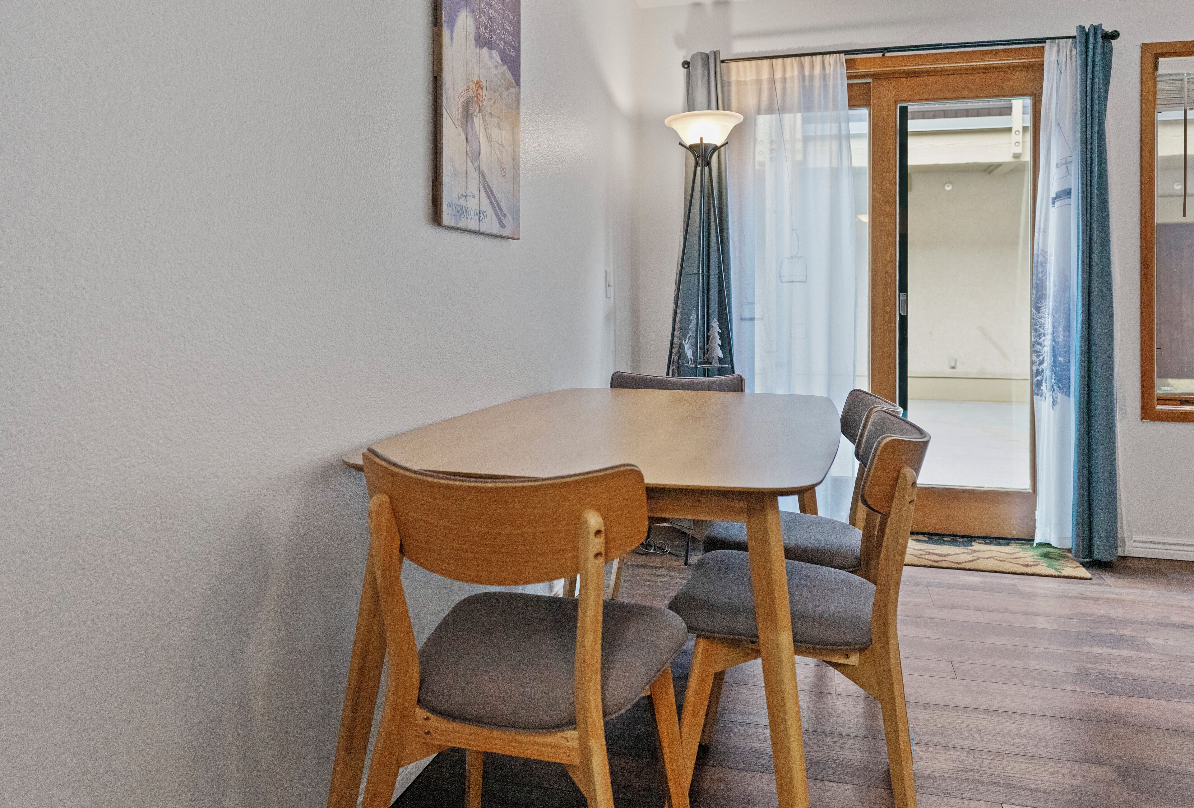 A dining area with a wooden table and four chairs, a floor lamp, and a sliding glass door with curtains.