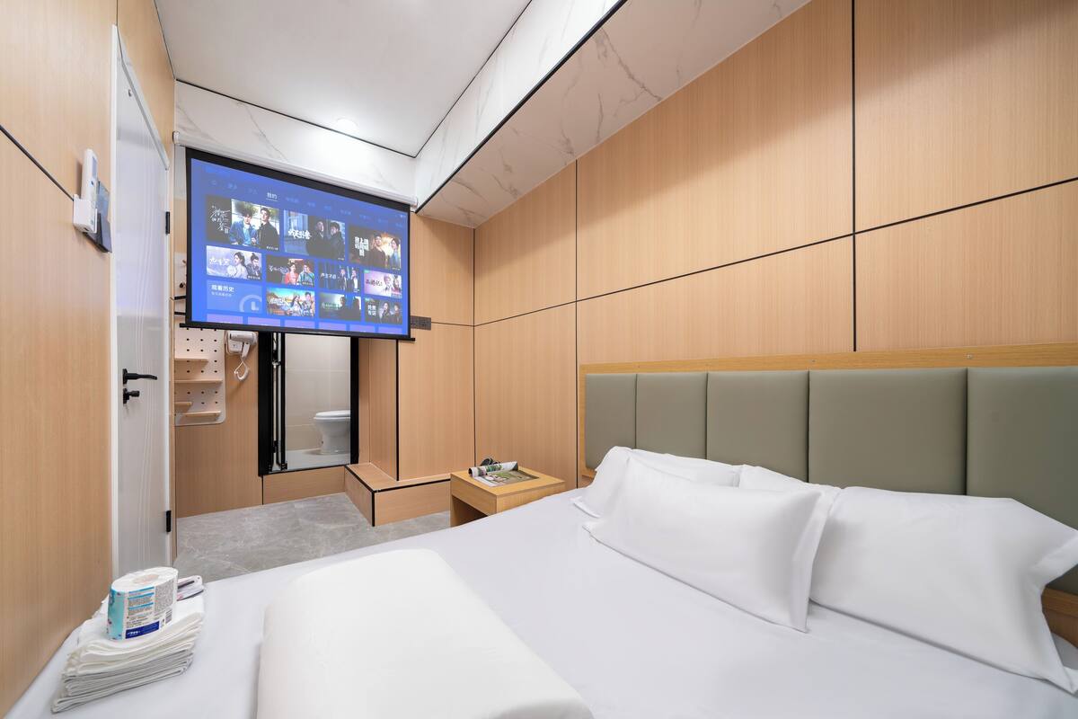 Habitación de hotel con una cama, un televisor montado en la pared, un armario de madera y una mesita de noche pequeña.