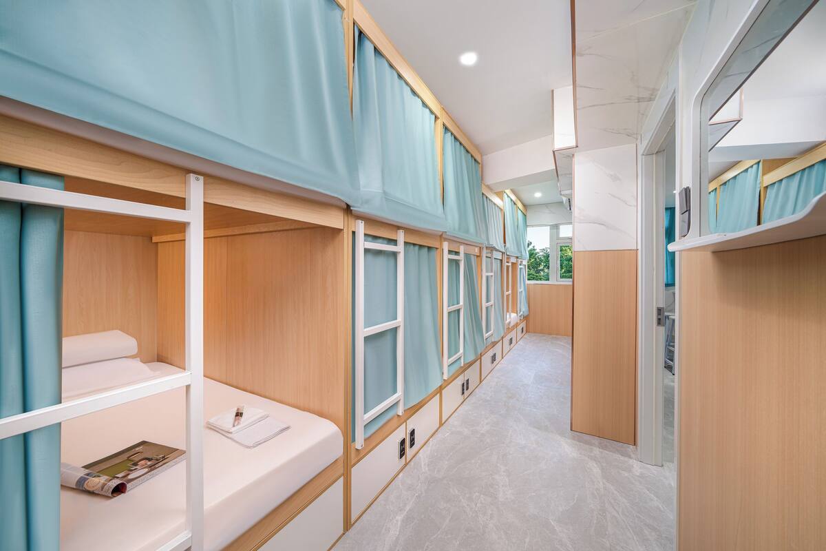 Una hilera de camas literas con estructura de madera y cortinas azules, un suelo de mármol y vistas al exterior con vegetación.