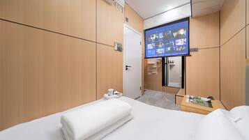 A hotel room with a bed, a TV mounted on the wall displaying a menu, a small table with a magazine, and a door leading to a bathroom.