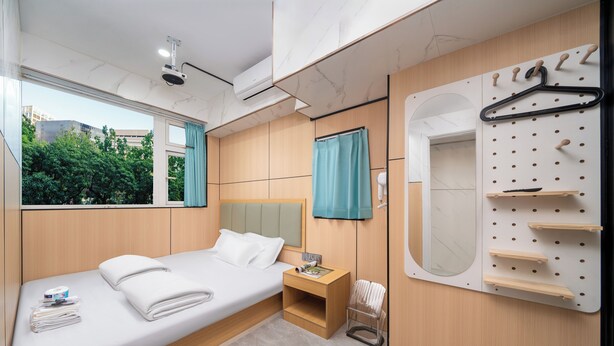 A modern hotel room with a bed, a nightstand, a mirror, a wall-mounted drying rack, and a view of greenery outside. - Windsor Hella Inn (Kowloon)