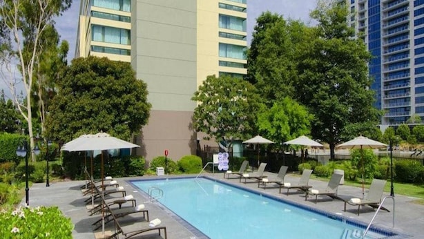 A swimming pool with lounge chairs and umbrellas, surrounded by greenery and tall buildings. - Orange County Hotel (Santa Ana)