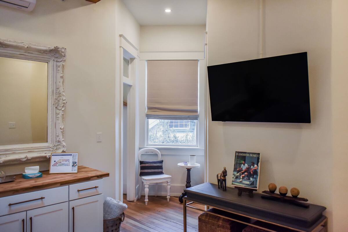 A room with a wooden vanity, a mirror, a television mounted on the wall, a small table with a decorative item, and a window with a view of the outside.