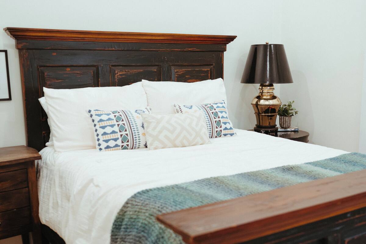 A neatly made bed with a patterned blanket, a wooden headboard, and a bedside table with a lamp and a small plant.