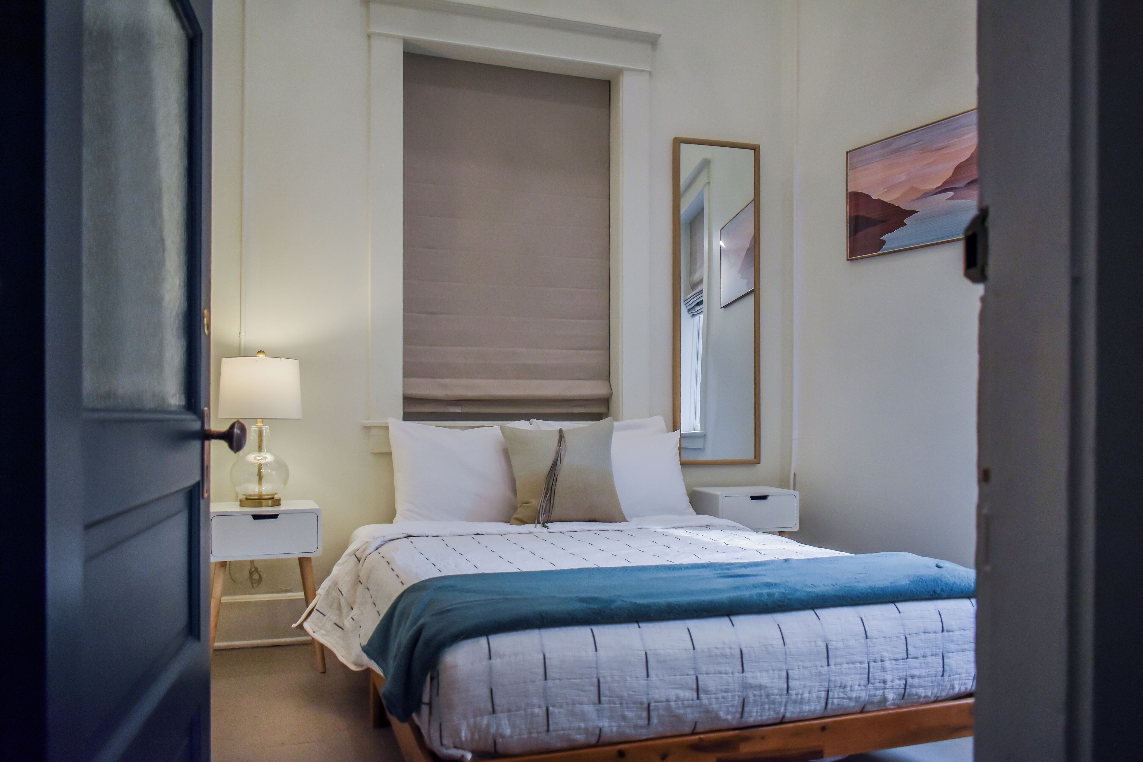 A bedroom with a bed, bedside tables, a lamp, a window with blinds, and a painting on the wall.