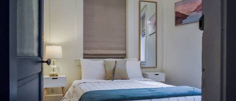 A bedroom with a bed, bedside tables, a lamp, a window with blinds, and a painting on the wall.
