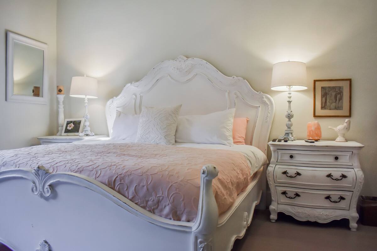 A white four-poster bed with a pink bedspread, two bedside tables with lamps, and a white dresser with drawers.