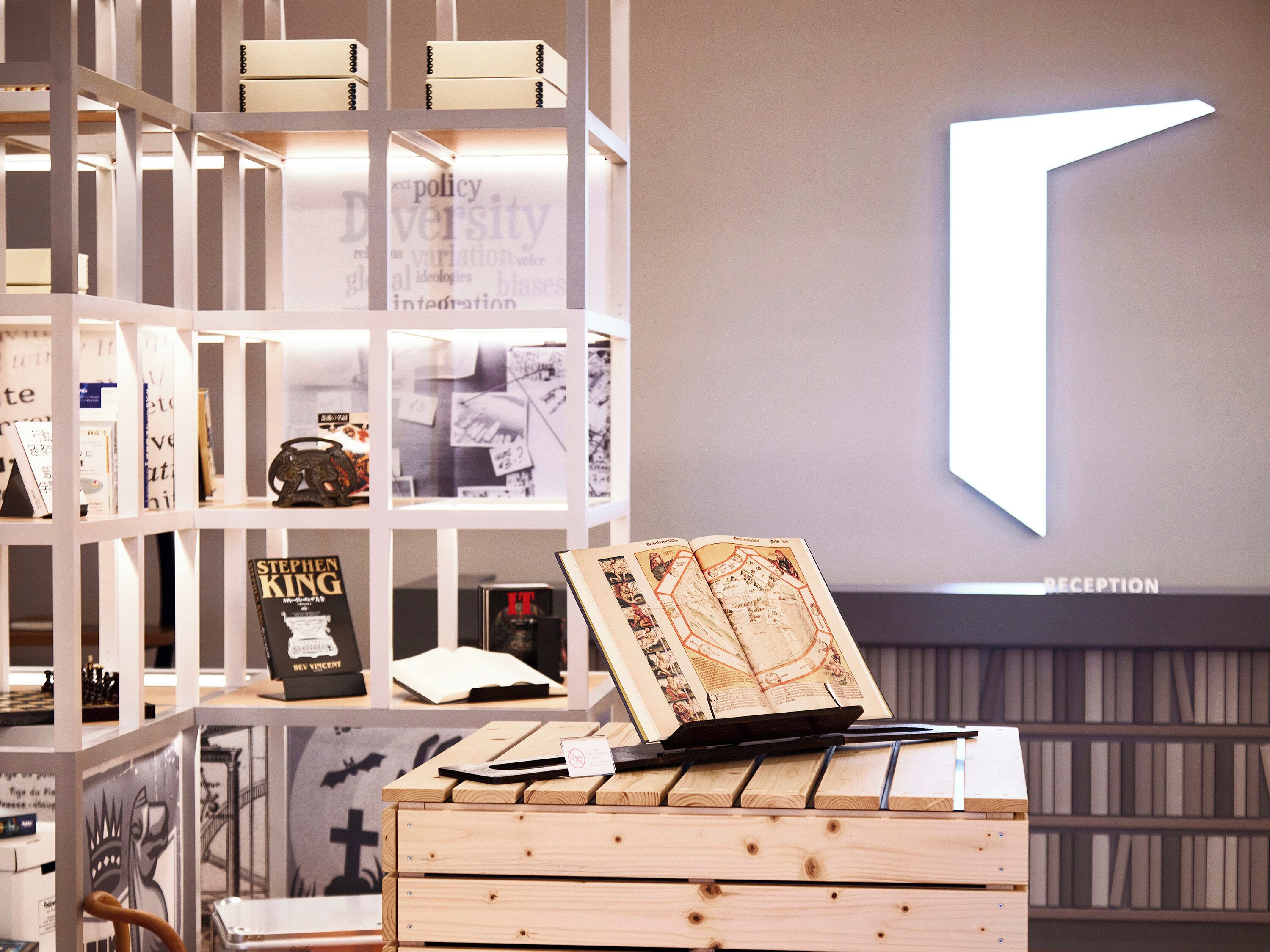 A reception area with a modern design, featuring a wooden desk with a laptop and a book, surrounded by shelves with books and a large illuminated 'T' sign.