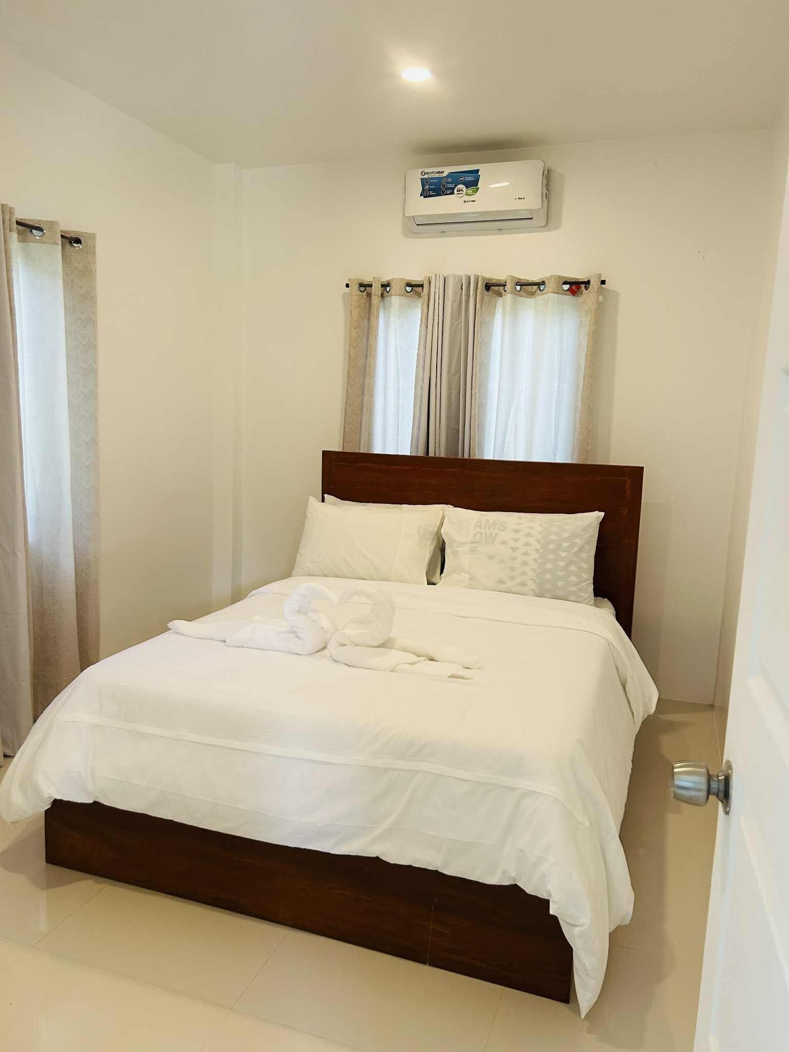 A neatly made bed with white linens and a wooden headboard, a white air conditioning unit above it, and a window with sheer curtains.