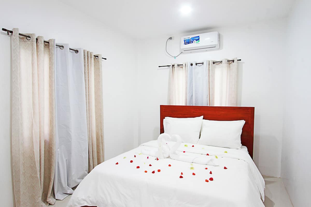 A hotel room with a bed, white bedding, a wooden headboard, and an air conditioning unit on the wall.