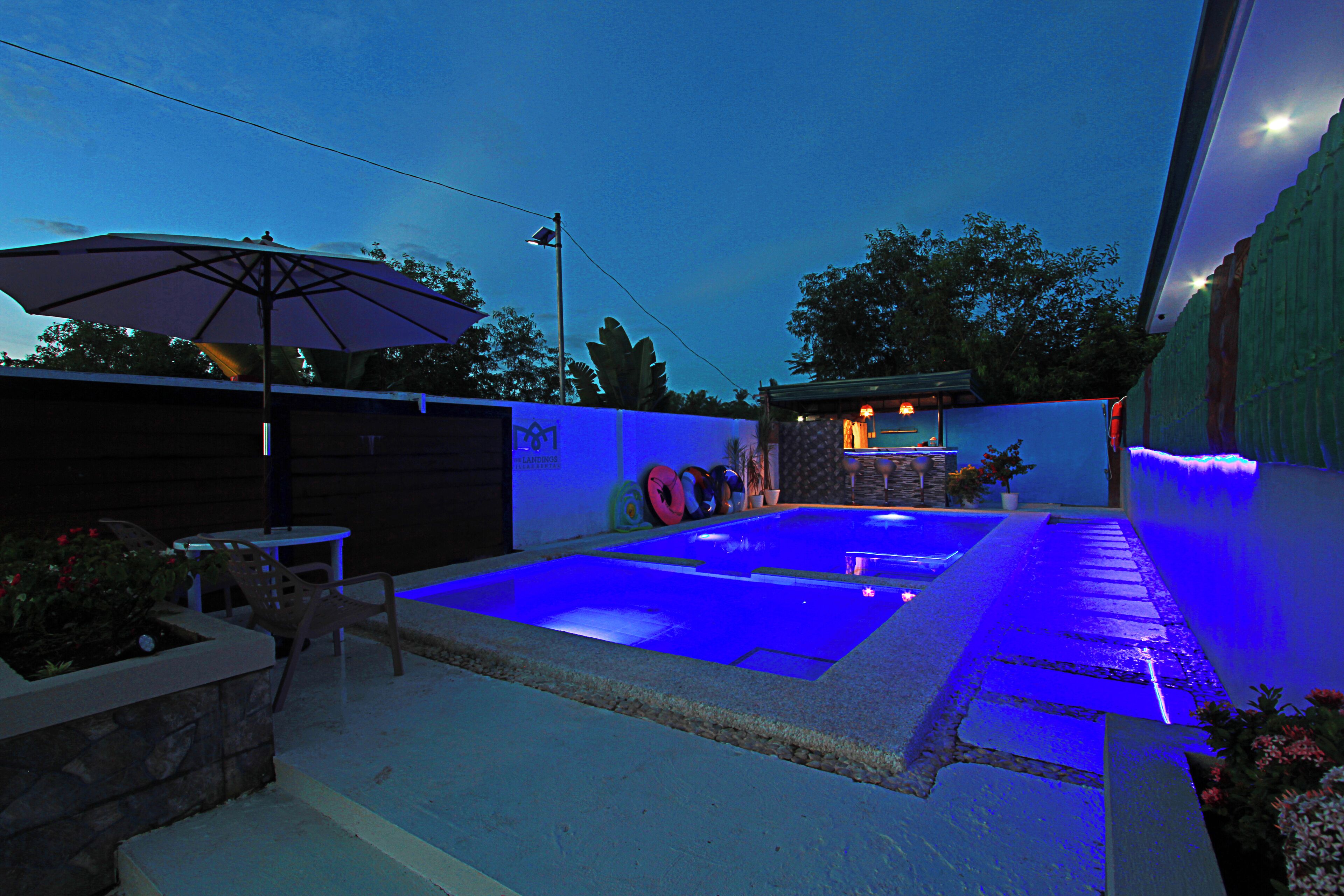 A swimming pool with blue lighting, surrounded by a stone wall and a covered patio area with an umbrella.