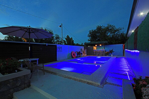 A swimming pool with blue lighting, surrounded by a stone wall and a covered patio area with an umbrella. - the Landing Villas Rentals (Panglao)