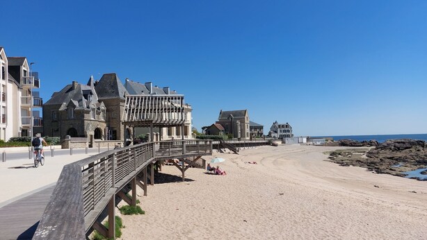 Un paseo marítimo en la playa con un camino de madera, edificios de piedra y un cielo azul despejado.