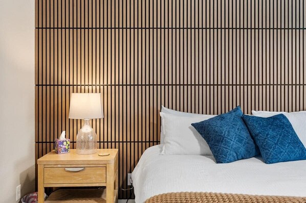 Quarto com cabeceira de ripas de madeira, mesa de cabeceira com abajur e duas almofadas azuis na cama.