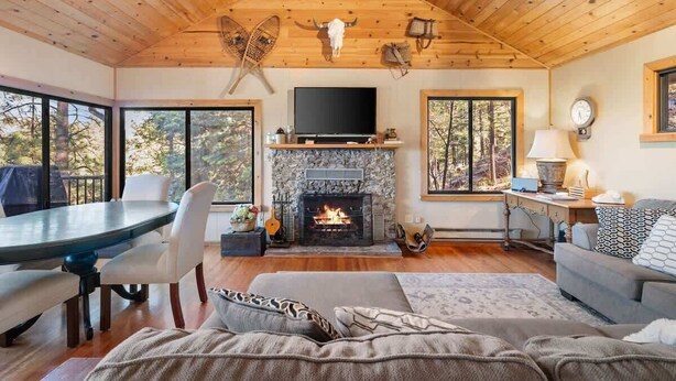 A cozy cabin interior with a stone fireplace, wooden ceiling, and large windows offering a forest view. - Prescott Pine Retreat | Views • Fireplace • Patio (Prescott)