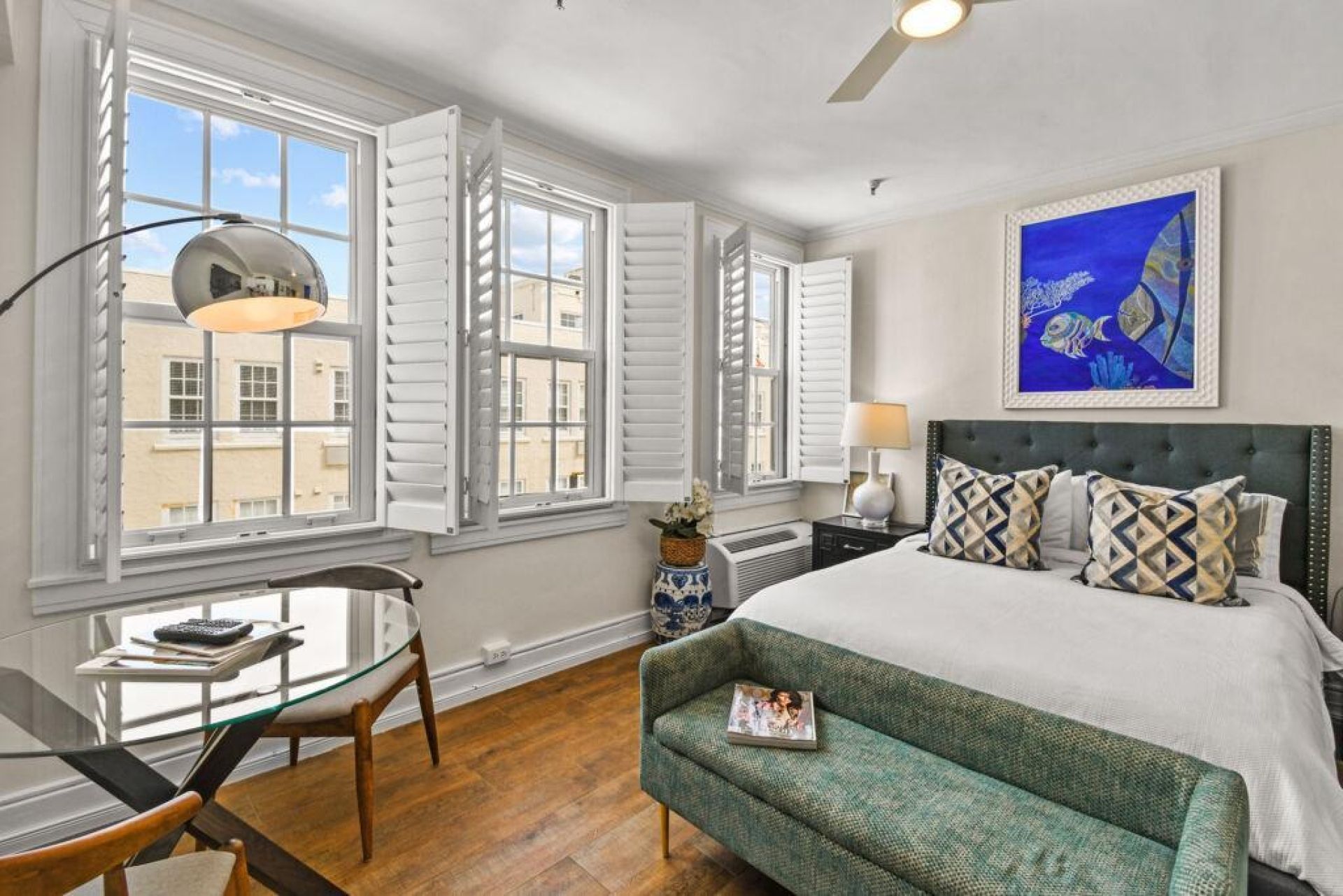 A hotel room with a bed, a desk, a chair, a sofa, and a large window with shutters.