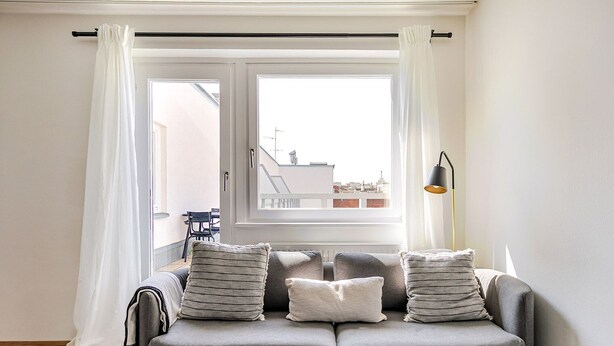 Un canapé gris avec des coussins à rayures, une fenêtre avec des rideaux blancs et un lampadaire noir.