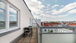 Un balcon avec une table et des chaises, donnant sur un paysage urbain avec des bâtiments aux toits rouges.