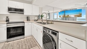 A modern kitchen with white cabinetry, a built-in oven, a dishwasher, and a large window with a beach view. - 1605 Oceanfront Pelican Beach Resort Amazing Location Multiple Pools & Hot Tubs (Destin)