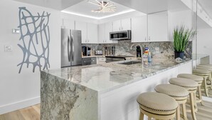 A modern kitchen with white cabinets, a large island with stools, a built-in microwave, and a unique ceiling light fixture. - 1605 Oceanfront Pelican Beach Resort Amazing Location Multiple Pools & Hot Tubs (Destin)