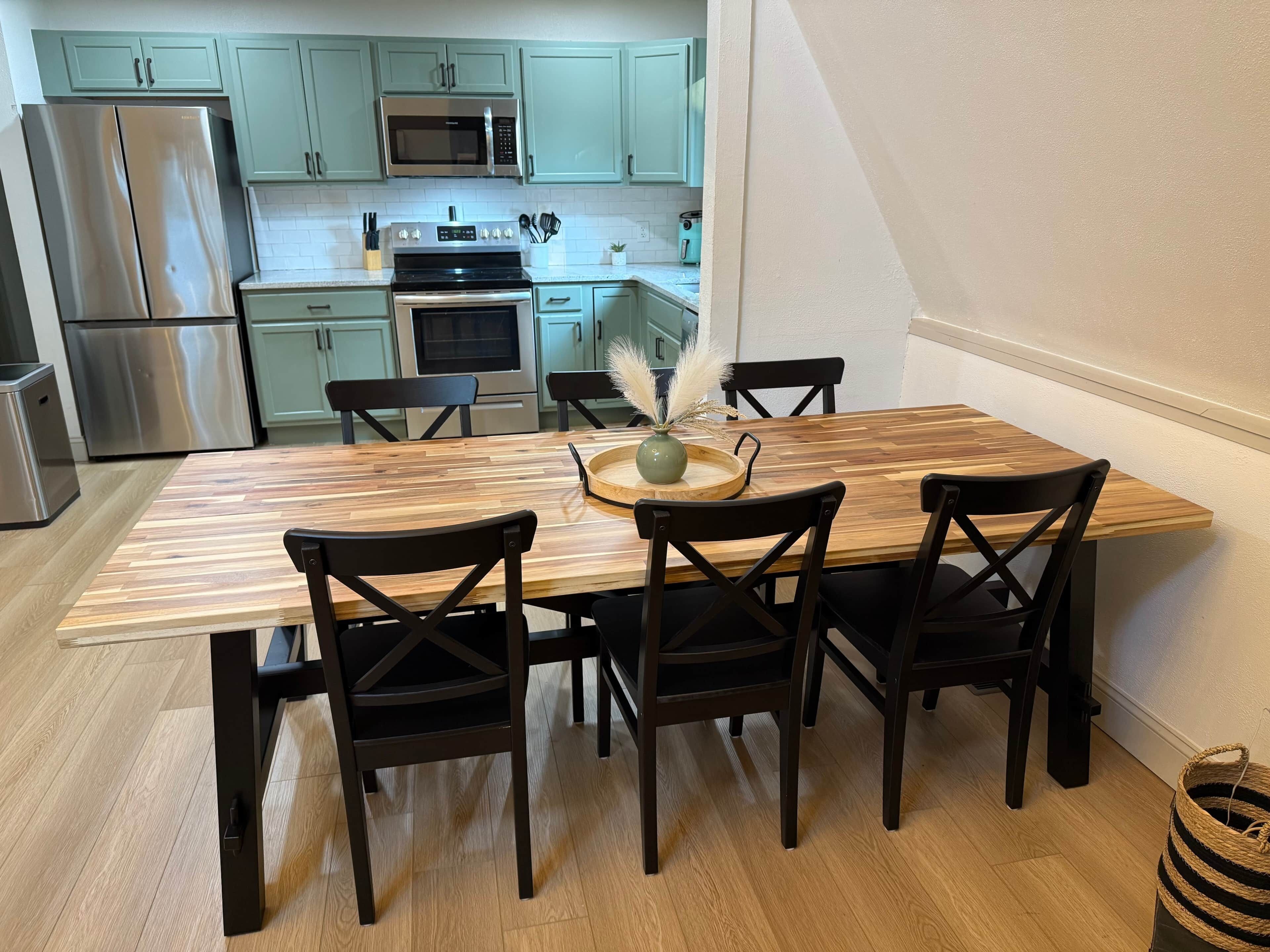 Un comedor con una mesa de madera y sillas negras, una cocina al fondo con armarios verde claro, y un pequeño florero decorativo sobre la mesa.