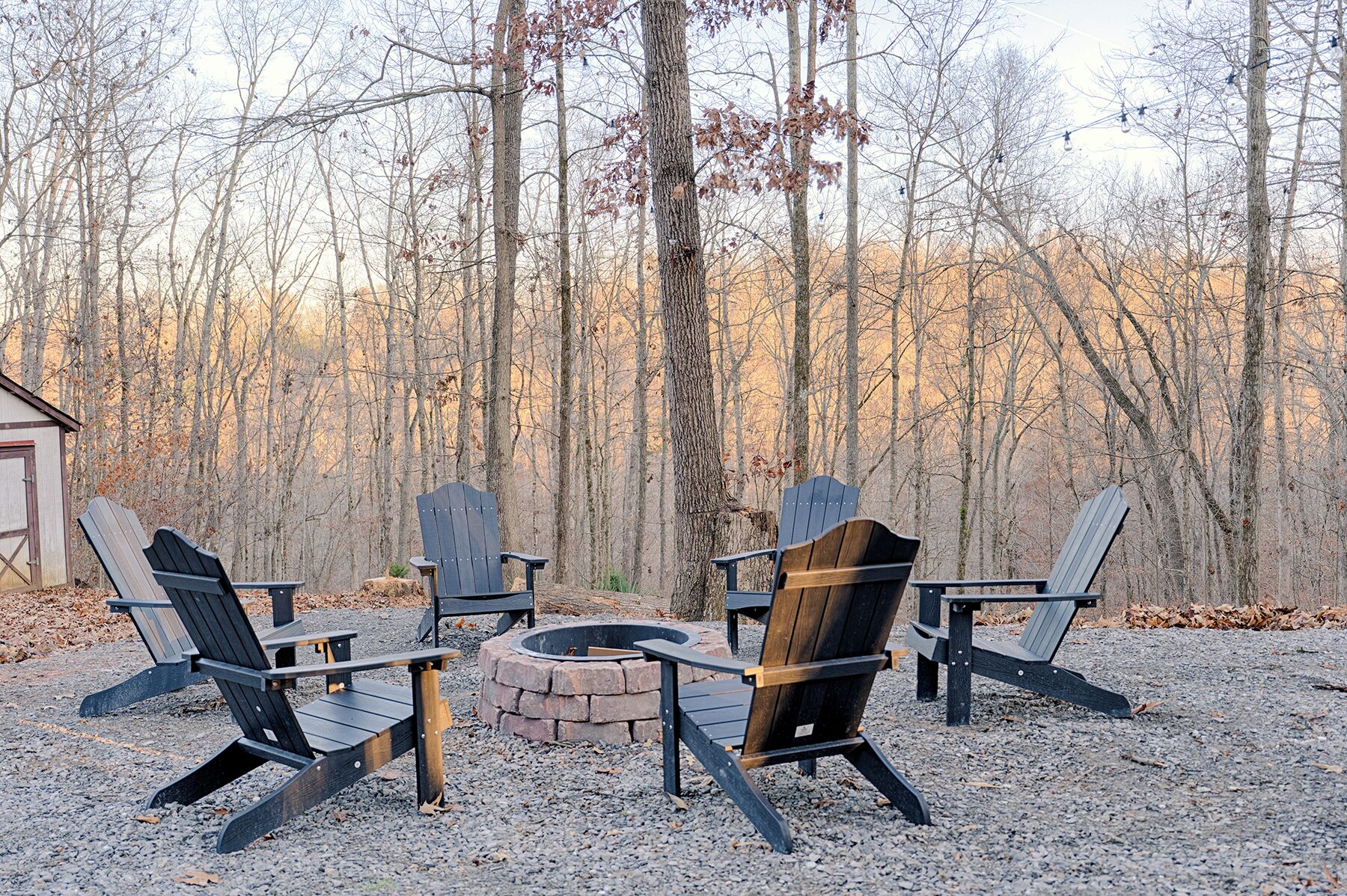 A circular fire pit surrounded by four Adirondack chairs in a forest setting.
