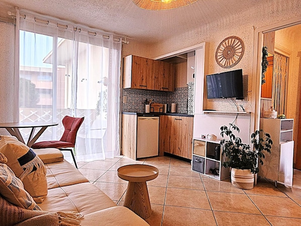 A modern living room with a sofa, a small table, and a kitchen area with wooden cabinets and a TV. - Beach 200 m • Terrace • Fréjus-Plage (Fréjus)