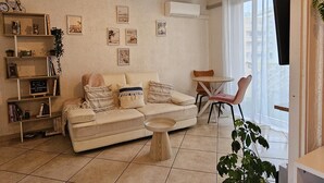 A living room with a beige sofa, a round wooden coffee table, and a small round table with a chair. There are wall-mounted shelves with decorative items and a few framed pictures. An air conditioning unit is mounted on the wall, and a window with curtains is visible. - Modern 30m² Apartment with A/C & Wi-Fi – Just 100m from the Beach (Fréjus)
