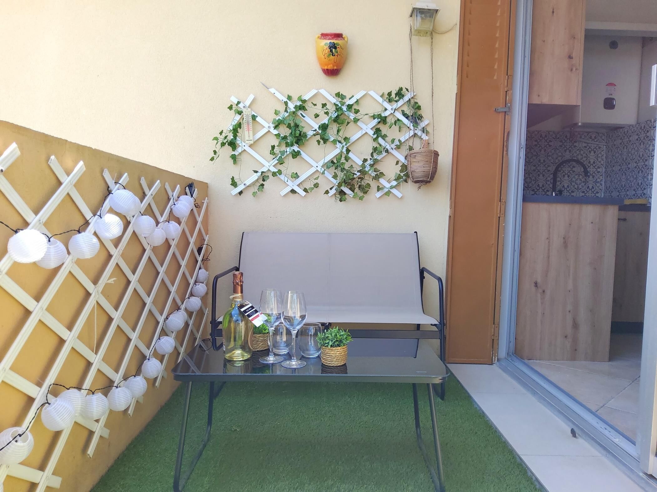 A glass table with two wine glasses and a bottle, a white bench, and a lattice wall with hanging lights and greenery.