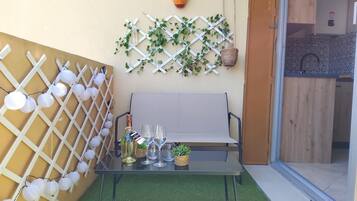 A glass table with two wine glasses and a bottle, a white bench, and a lattice wall with hanging lights and greenery.