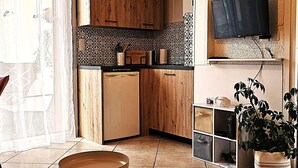A compact kitchen with wooden cabinets, a built-in dishwasher, a tiled backsplash, a flat-screen TV mounted above the counter, and a potted plant. - Modern 30m² Apartment with A/C & Wi-Fi – Just 100m from the Beach (Fréjus)