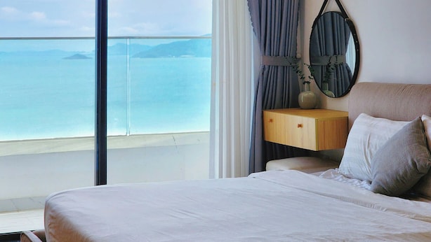 A bedroom with a large window overlooking a body of water, a bed with pillows, a bedside table, and a mirror on the wall. - TMS QUY NHON- RUBY LUXURY SEA APARTMENT (Quy Nhon)