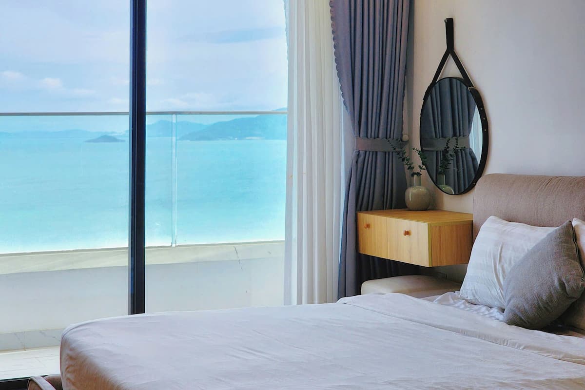A bedroom with a large window overlooking a body of water, a bed with pillows, a bedside table, and a mirror on the wall.